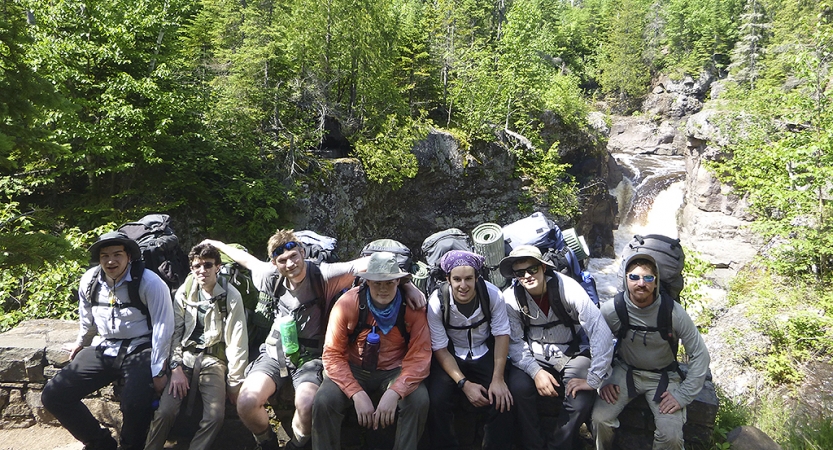 A group of people wearing backpacks sit on a rocky overlook above trees